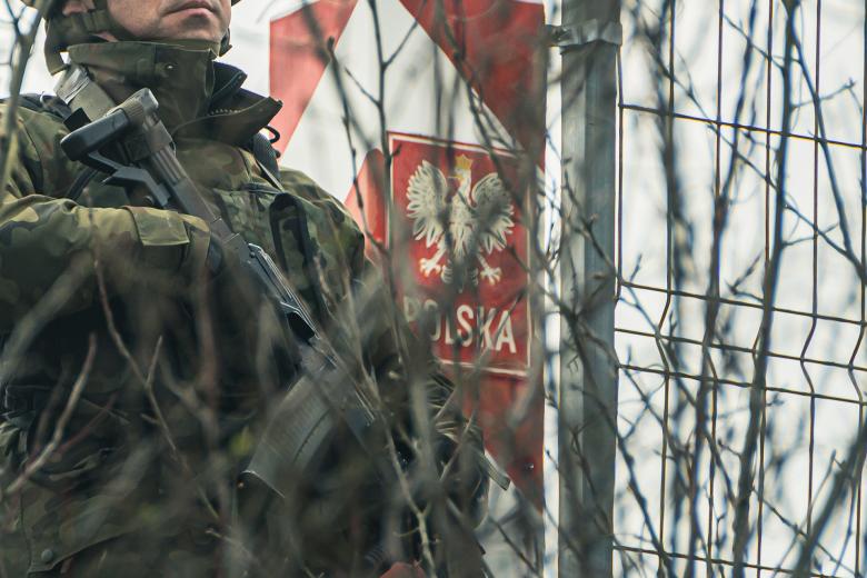 Żołnierz trzymający karabin stoi przy biało-czerwonym słupie granicznym z herbem Polski i napisem "Polska"