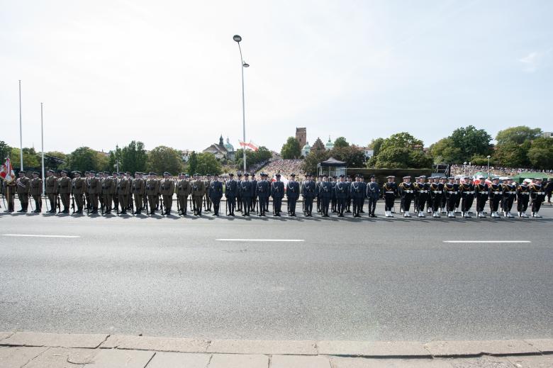 Żołnierze biorący udział w defiladzie, w mundurach reprezentujących poszczególne rodzaje sił zbrojnych, stoją na baczność przed publicznością obserwującą defiladę