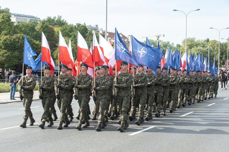 Maszerująca w defiladzie kolumna żołnierzy, którzy trzymają flagi Polski, Unii Europejskiej i NATO