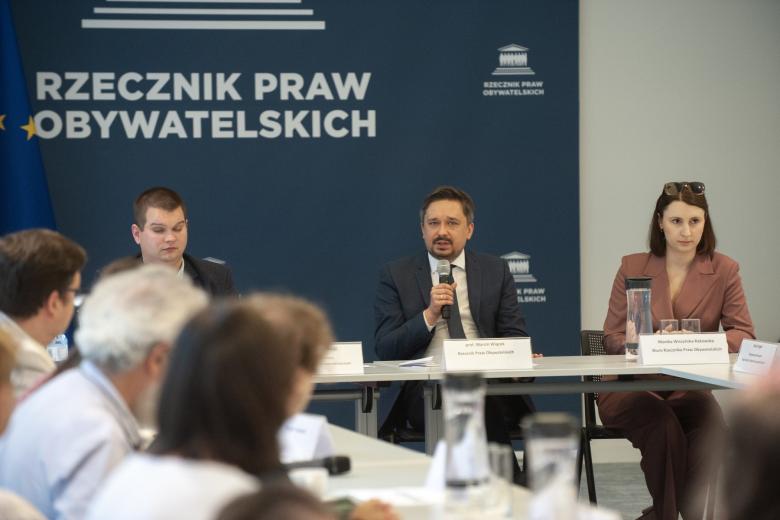 Trzy osoby siedzą za stołem w sali konferencyjnej, za nimi granatowa plansza z napisem "Rzecznik Praw Obywatelskich"