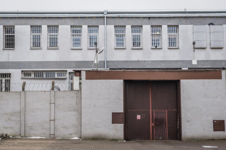 Stalowa brama zakładu karnego w wysokim murze zakończonym drutem kolczastym. Za bramą widoczny jest budynek z okratowanymi oknami
