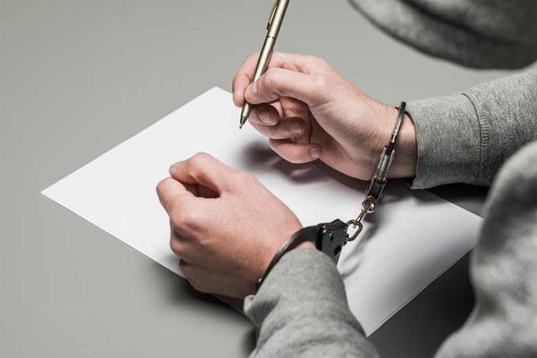 Mężczyzna w kajdankach z długopisem w dłoni pochyla się nad kartką papieru