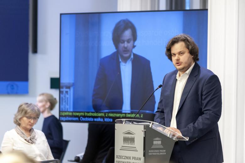 Osoba przemawia za mównicą na tle wyświetlacza z relacją internetową z konferencji