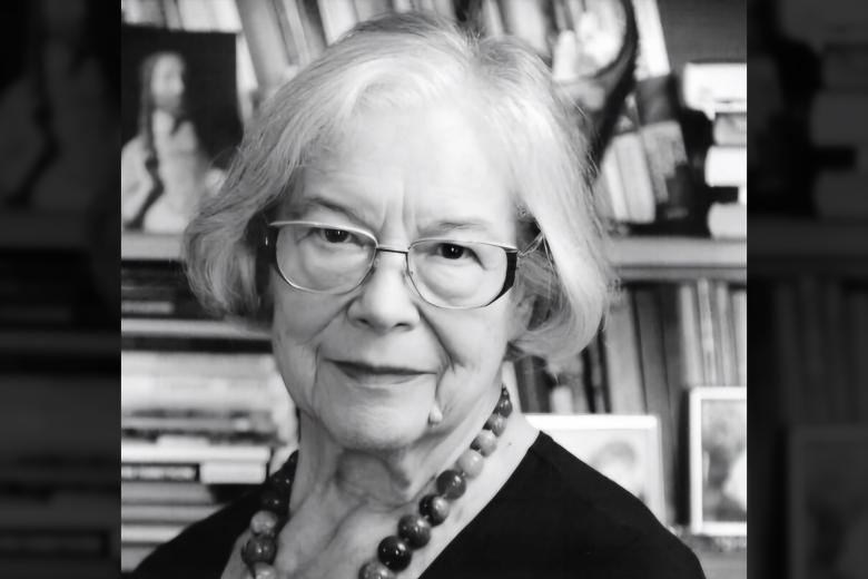 Czarno-biały portret Krystyny Mrugalskiej - kobieta w okularach i z koralami na szyi na tle regału z książkami i zdjęciami