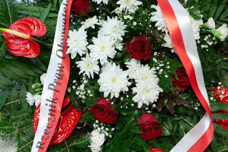 Zielony wieniec z białymi i czerwonymi kwiatami i biało-czerwoną wstążką z napisem Rzecznik Praw Obywatelskich