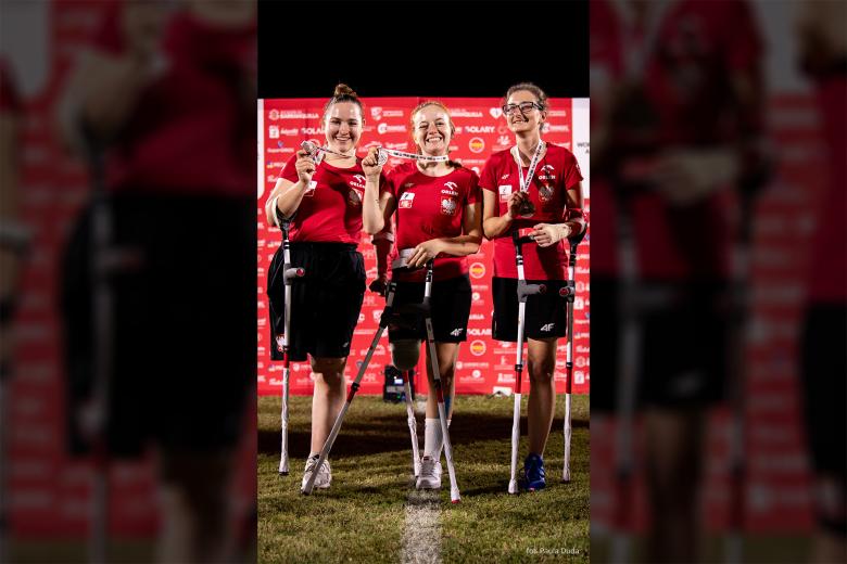 Trzy kobiety, każda z amputowaną jedną z nóg i wspierająca się na kulach, uśmiechają się pozując do zdjęcia z medalami w dłoniach