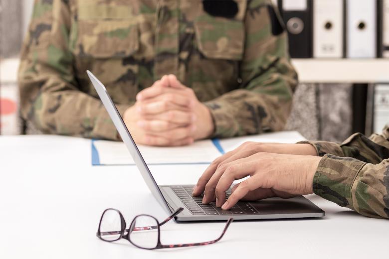 Dwie osoby w mundurach wojskowych siedzą przy stole. Jedna z nich korzysta z laptopa, przy którym leżą okulary korekcyjne.