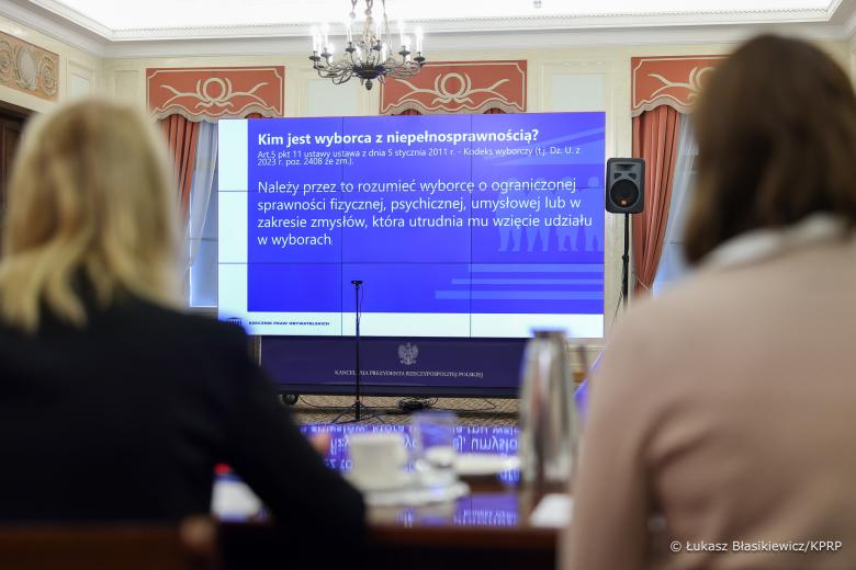 Duży ekran wyświetlacza z prezentacją, której przyglądają się osoby na sali konferencyjnej