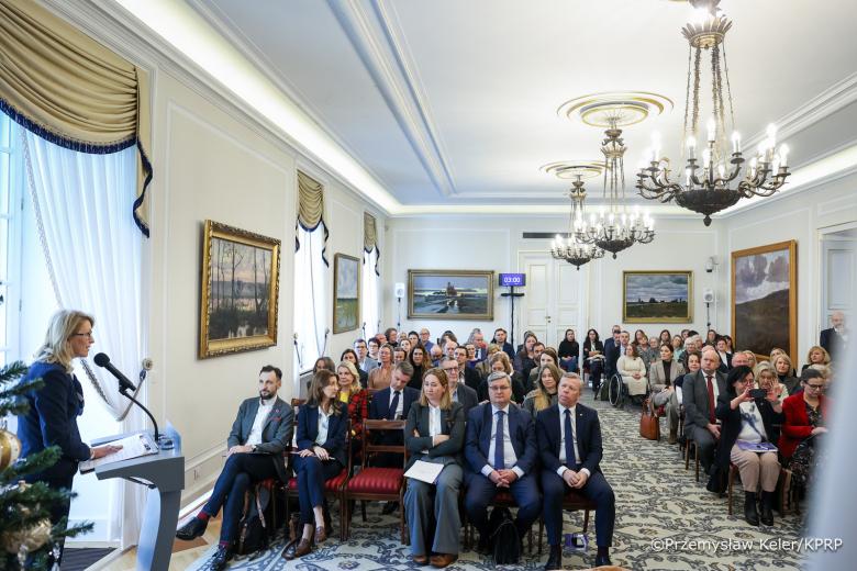 Kilkadziesiąt osób siedzi w zabytkowej sali i słucha wystąpienia osoby mówiącej za mównicą