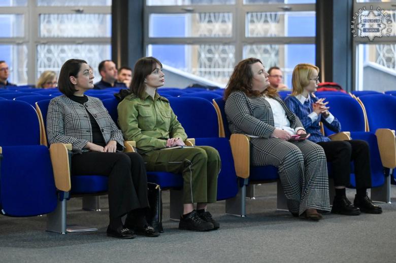 Cztery osoby siedzą w pierwszym rzędzie na dużej sali wykładowej i słuchają wystąpienia