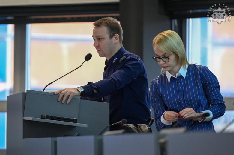 Osoba w mundurze przemawia za mównicą, obok druga osoba przegląda notatki przygotowując się do wystapienia