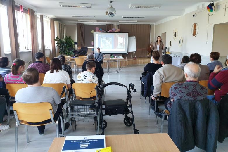 Kilkanaście osób siedzących na krzesłach słucha wystąpienia