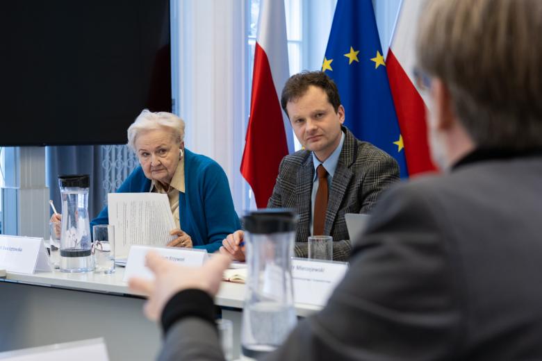 Dwie osoby siedzą przy stole i z uwagą kogoś słuchają