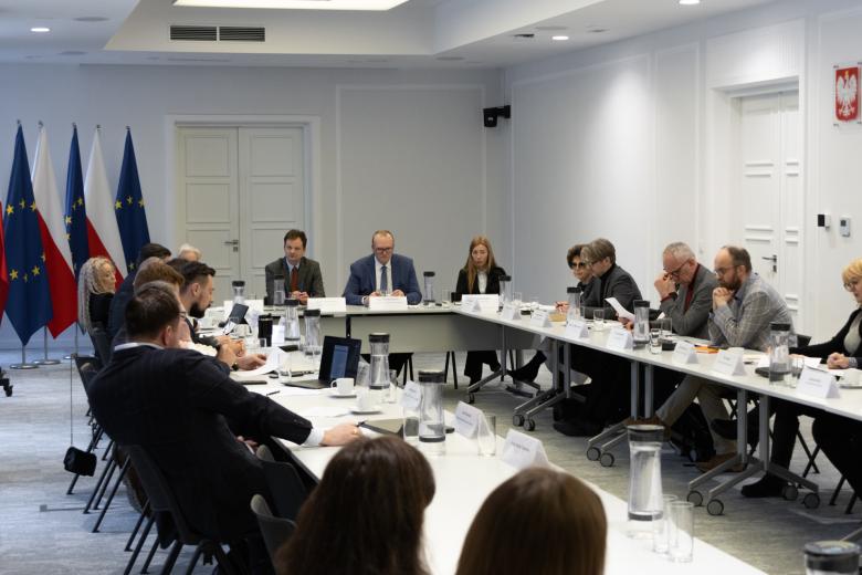 Kilkadziesiąt osób siedzi przy stołach ustawionych w podkowę na dużej sali konferencyjnej