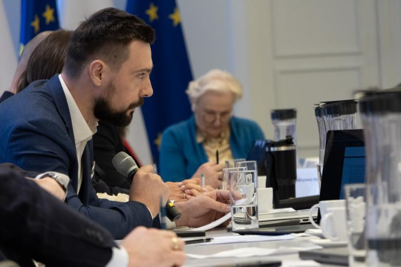 Osoby siedzą przy stole, jedna mówi do mikrofonu trzymanego w dłoni