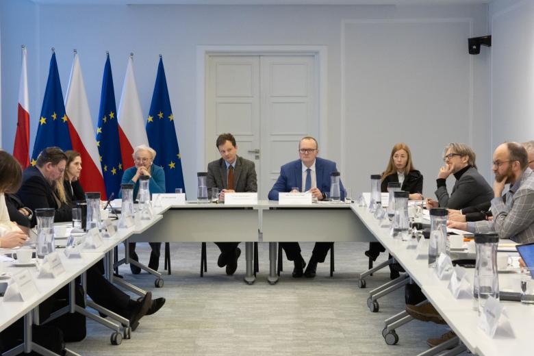 Kilkanaście osób siedzi przy stołach ustawionych w podkowę na sali konferencyjnej