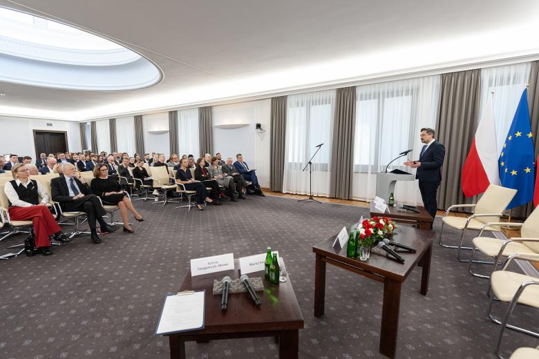 Osoba przemawia za mównicą, przed nią kilkadziesiąt osób siedzi na krzesłach na sali konferencyjnej i słucha