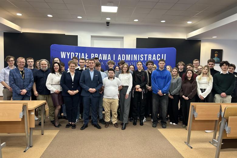 Kilkadziesiąt osób pozuje na stojąco do wspólnego zdjęcia w sali wykładowej