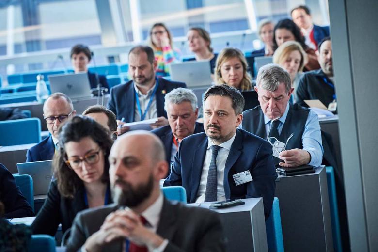 Osoby siedzą w ławach na dużej sali konferencyjnej