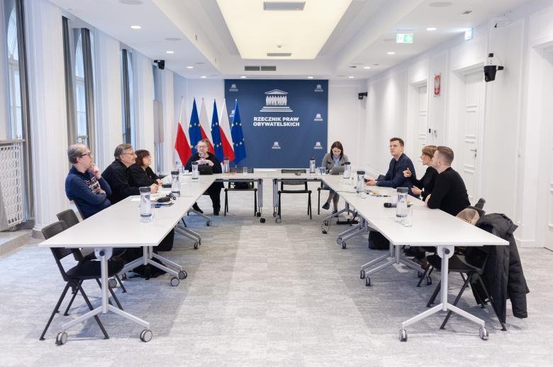 Kilka osób siedzi przy stołach ustawionych w podkowę na sali konferencyjnej