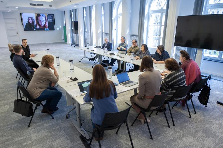 Kilkanaście osób rozmawia, siedząc przy stole konferencyjnym o kształcie podkowy. W szczycie stołu znajduje się ekran, na którym wyświetlane są osoby uczestniczące w spotkaniu za pośrednictwem internetu.