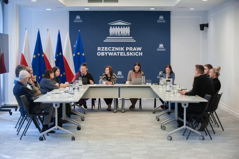 Kilkanaście osób rozmawia, siedząc przy stole konferencyjnym o kształcie podkowy.