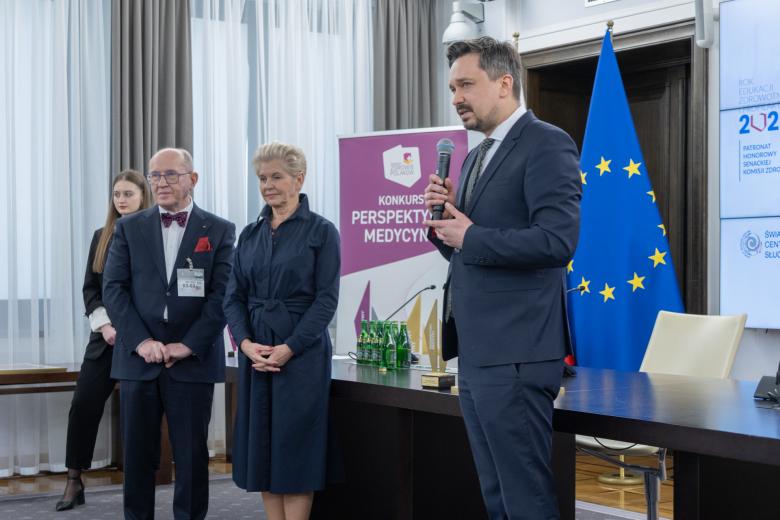 Marcin Wiącek wypowiada się do mikrofonu stojąc przed publicznością. Obok niego stoją trzy osoby.