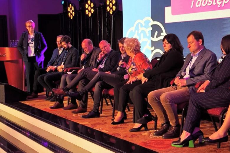 Kilkanaście osób siedzi na krzesłach w rzędzie na scenie, jedna z nich mówi do mikrofonu