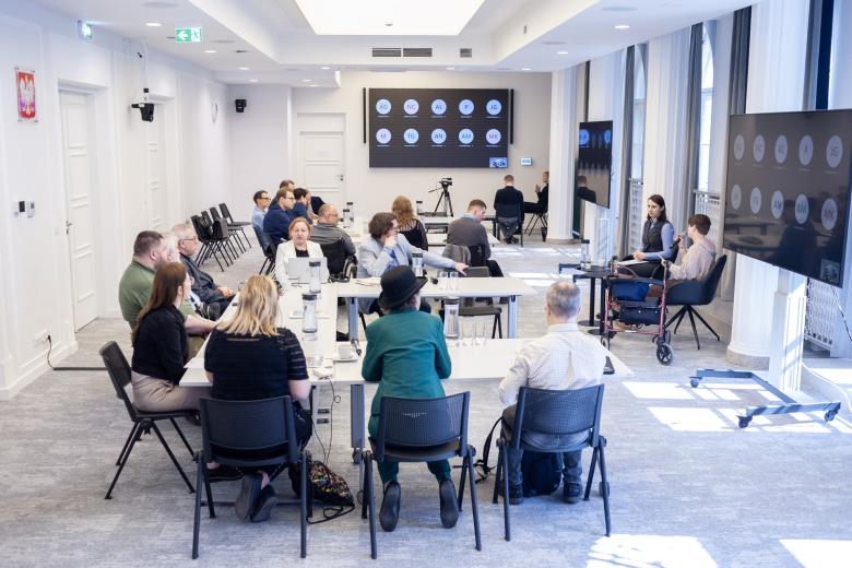 Kilkadziesiąt osób siedzi przy stołach na sali konferencyjnej, na dużych ekranach widoczni uczestnicy spotkania łączący się za pomocą oprogramowania do komunikowania się na odległość