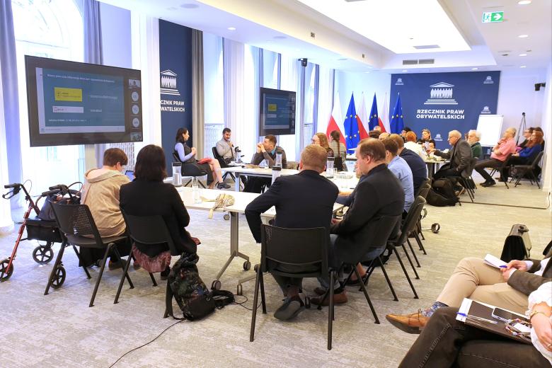 Kilkadziesiąt osób siedzi na sali konferencyjnej i słucha wystąpienia dwóch osób siedzących na krzesłach na środku