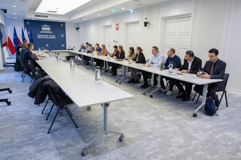 Kilkanaście osób siedzi przy stołach ustawionych w podkowę, za stołami przy ścianie rząd flag Polski i UE i duża plansza z białym napisem Rzecznik Praw Obywatelskich na granatowym tle