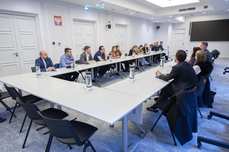 Kilkanaście osób siedzi przy stołach ustawionych w podkowę na dużej sali konferencyjnej