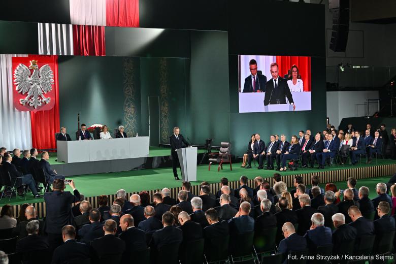 Kilkadziesiąt osób siedzących w dużej hali słucha wystąpienia osoby mówiącej za mównicą, na ścianie w tyle duże godło Polski - biały orzeł w koronie na czerwonym polu