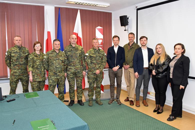 Dziesięć osób, wśród których 5 jest umundurowanych, pozuje do zdjęcia w sali konferencyjnej. Za pozującymi osobami stoją flagi Polski i Unii Europejskiej oraz makieta słupa granicznego.