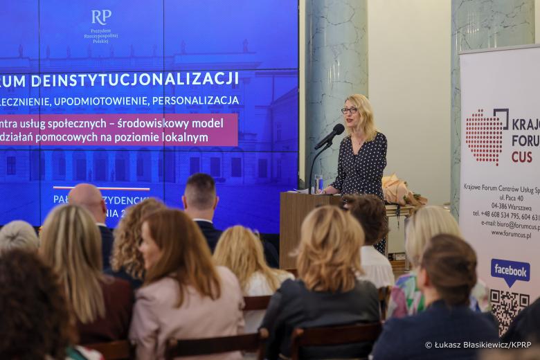 Osoba za mównicą przemawia do kilkudziesięciu osób siedzących na krzesłach na sali i słuchających