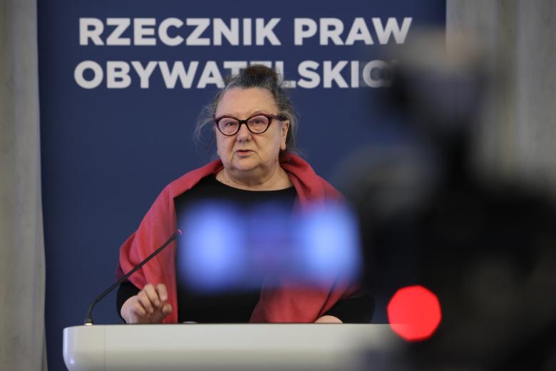 Barbara Imiołczyk wypowiada się do mikrofonu stojąc przy mównicy. Na pierwszym, rozmytym planie widać skierowaną w jej stronę kamerę wideo.
