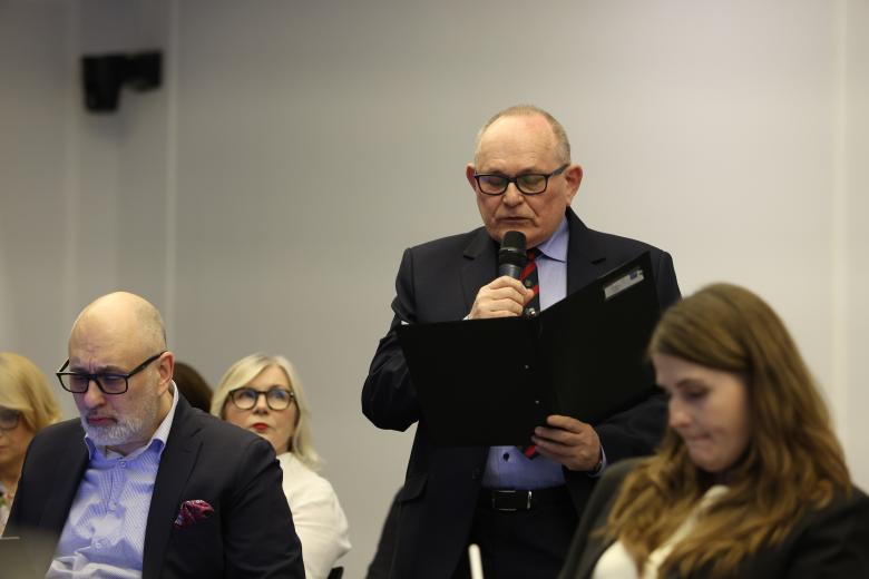 Mężczyzna stoi i wypowiada się do mikrofonu, trzymając folder z dokumentami. Obok niego siedzą inni uczestnicy debaty