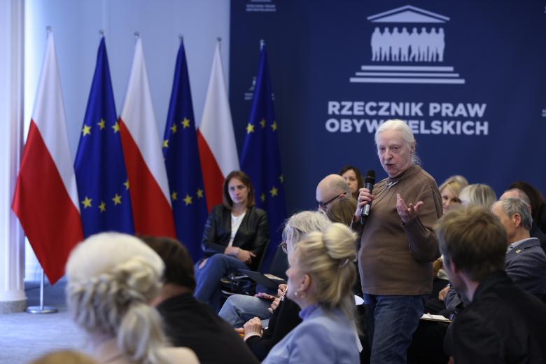 Uczestniczka debaty wypowiada się na stojąco do mikrofonu, gestykulując. Wokół niej siedzą inni uczestnicy debaty.