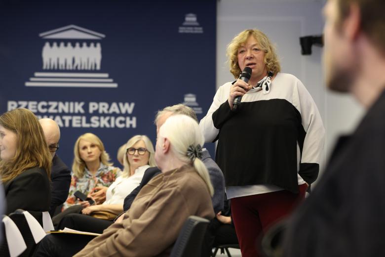 Kobieta stoi i wypowiada się do mikrofonu. Wokół niej siedzą inni uczestnicy debaty.