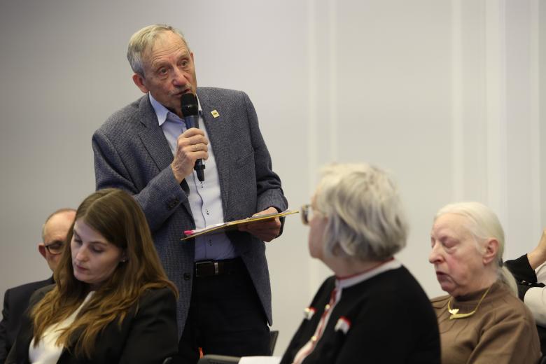 Mężczyzna wypowiada się do mikrofonu stojąc i trzymając dokumenty. Wokół niego siedzą inni uczestnicy debaty.