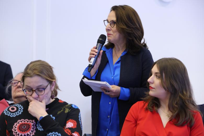 Kobieta wypowiada się do mikrofonu, trzymając notes. Wokół niej siedzą inne osoby uczestniczące w debacie.