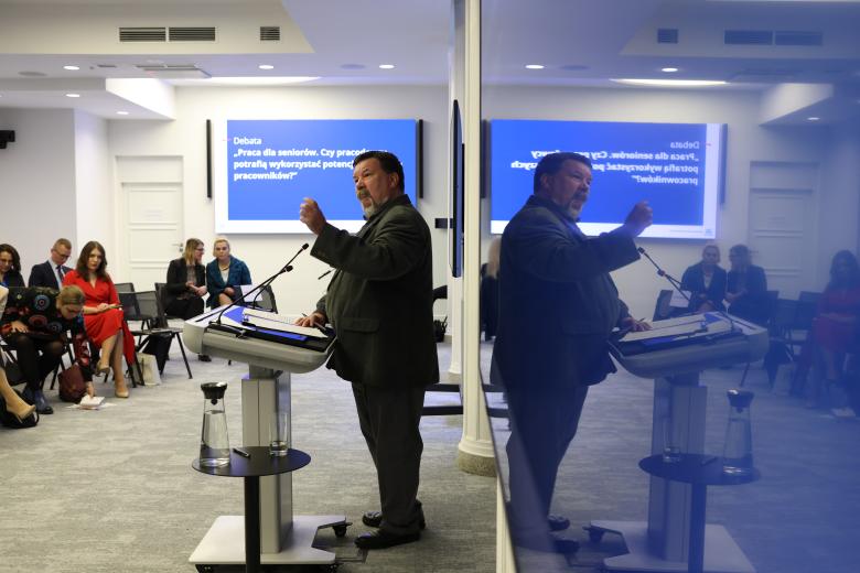 Tomasz Schimanek wypowiada się przy mównicy, gestykulując. Przed nim w kilku rzędach siedzą inni uczestnicy wydarzenia. Na ścianie w tle wisi wyświetlacz, na którym jest nazwa debaty.