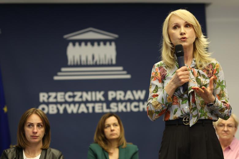 Uczestniczka debaty wypowiada się na stojąco do mikrofonu, gestykulując. Za nią siedzą inne osoby uczestniczące w debacie. W tle znajduje się ścianka z dużym logiem Rzecznika Praw Obywatelskich.