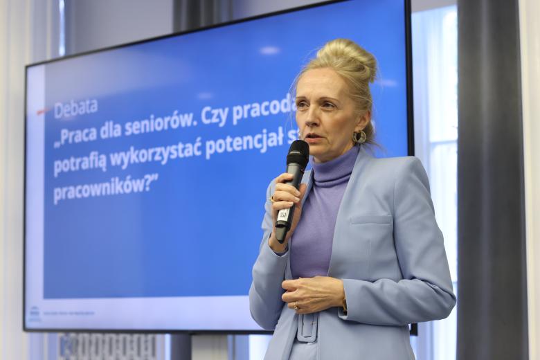 Kobieta stoi i wypowiada się do mikrofonu. Za nią znajduje się ekran wyświetlający nazwę debaty.