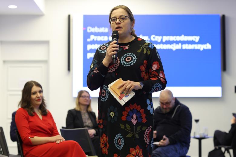 Kobieta stoi i wypowiada się do mikrofonu, trzymając w dłoni notes. Za nią siedzą inni uczestnicy debaty. W tle na ścianie jest ekran wyświetlający nazwę debaty.