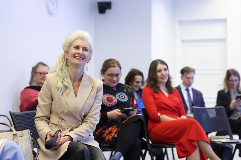 Kobieta siedzi i uśmiecha się, trzymając mikrofon. Obok niej i za nią siedzą inni uczestnicy debaty.