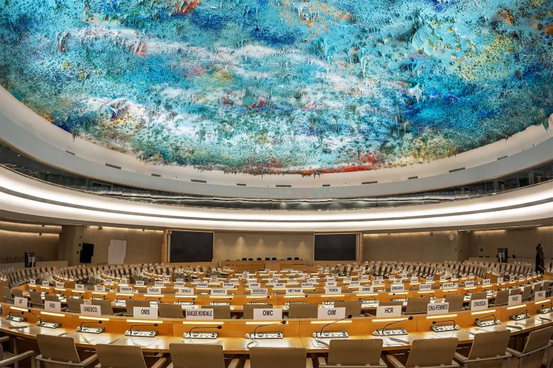 Sala obrad Rady Praw Człowieka ONZ. Kilka szeregów długich stołów jest ustawionych w okręgu wokół stołu prezydium. Nad stołami widać rzeźbę stanowiącą sufit pomieszczenia - kompozycję olbrzymiej ilości kolorowych, zastygniętych stalaktytów zwisających z sufitu.