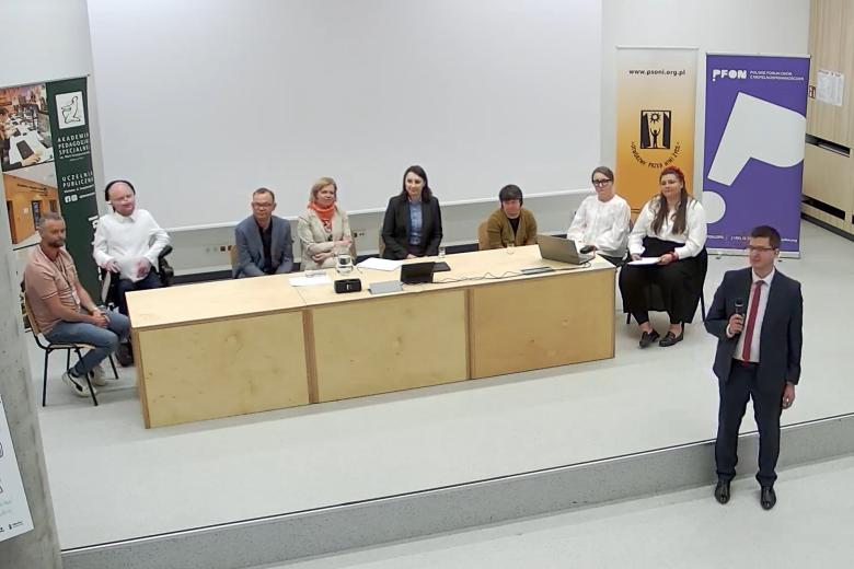 Osiem osób siedzi za długim stołem na podwyższeniu w sali wykładowej, obok mężczyzna stoi i mówi do mikrofonu