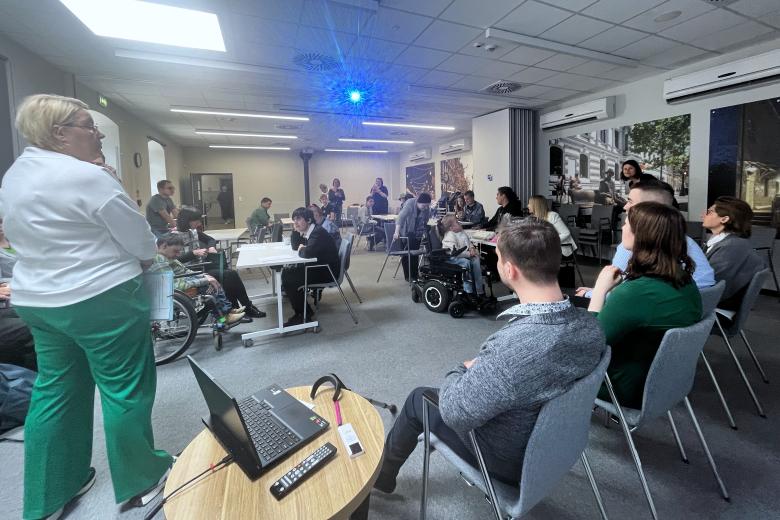 Kilkadziesiąt osób siedzi przy stołach na sali i słucha wystąpienia