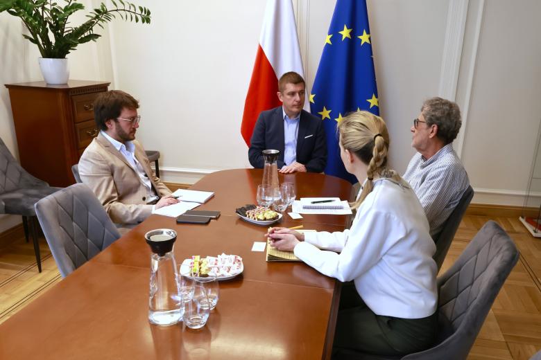 Cztery osoby siedzą przy stole i rozmawiają, w tle flagi Polski i UE